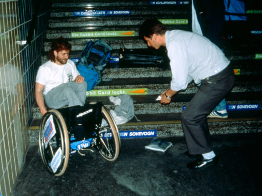 [1990] Paris? Usikkert sted. Hebbe har mistet rullestolen ned trappa og det ligger en del ting omkring ham og rullestolen. En mann hjelper til.