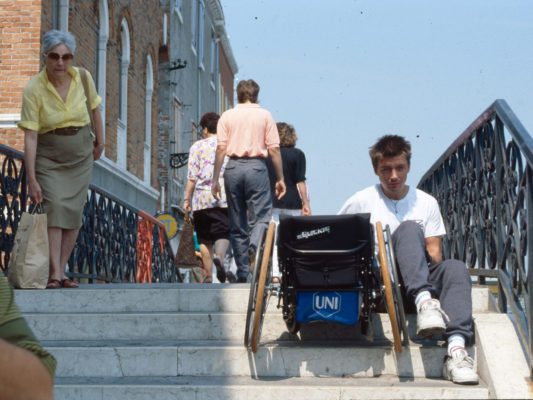 [1990] Venezia: Leif på veg opp trapp. Sitter på nest øverste trinn, trekker rullestolen etter seg. Ser tydelig sliten ut.