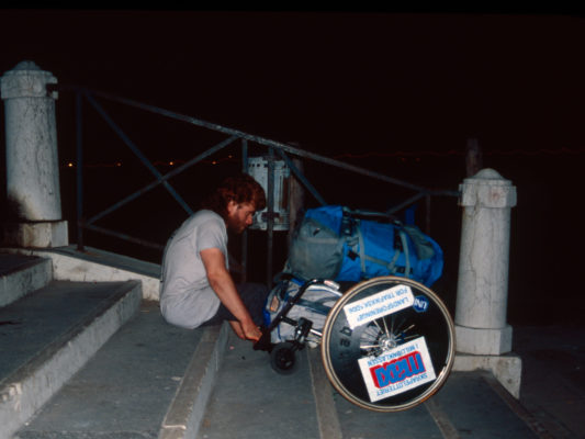 [1990] Venezia. Hebbe sitter i trapp og trekker rullestolen etter seg. Under setet på rullestolen er det en bag, og oppå rullestolen ligger en full ryggsekk. Det er mørkt.