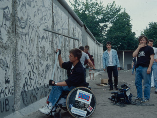 [1990] Leif, sittende i rullestol, hakker løs på Berlinmuren med en stor metall "T"..