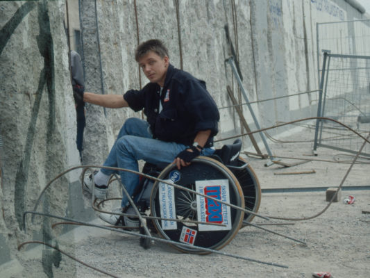 [1990] Leif i rullestol ved et hull i Berlinmuren. Mye bøyd armeringsjern omkring. Tydelig at muren er i ferd med plukkes fra hverandre.