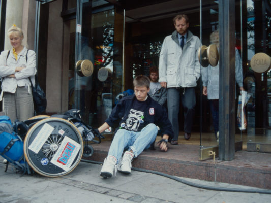 [1990] Leif sitter på trinn som fører inn til hovedinngangen til kjøpesenter. Trekker rullestolen etter seg.